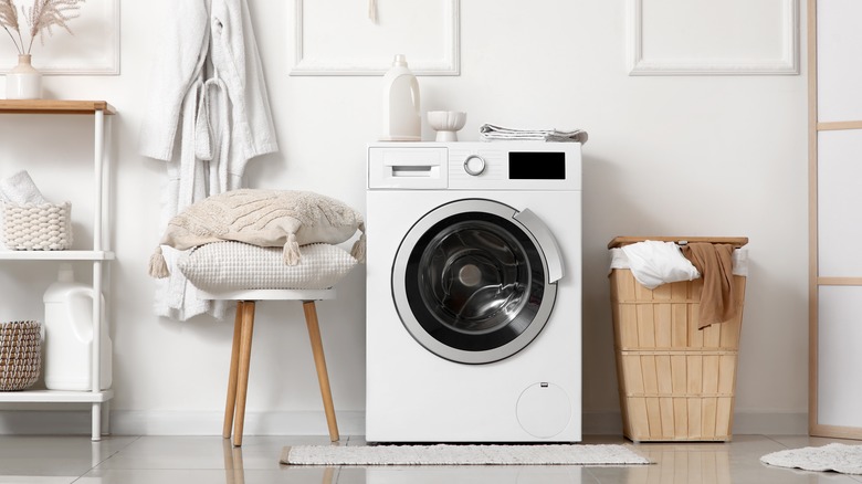 Modern washing machine with laundry basket and pillows on stool near white wall in bathroom