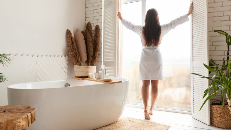 woman opening curtains with freestanding tub in bathroom