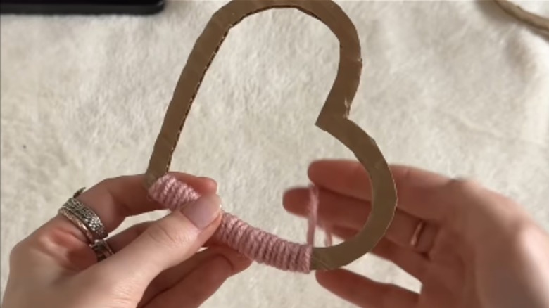 A pair of hands wrap a cardboard heart cutout with pink yarn.