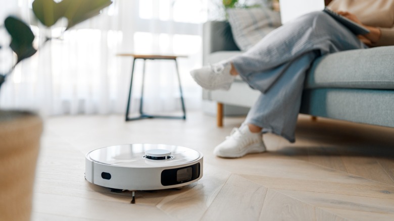 Robot vacuum cleaning hardwood floor in living room as person sits on sofa