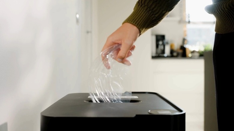 A hand inserts plastic packaging into a Clear Drop trash compactor.