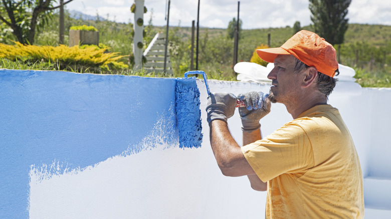 A man in an orange hat and yellow shirt paints a white pool blue