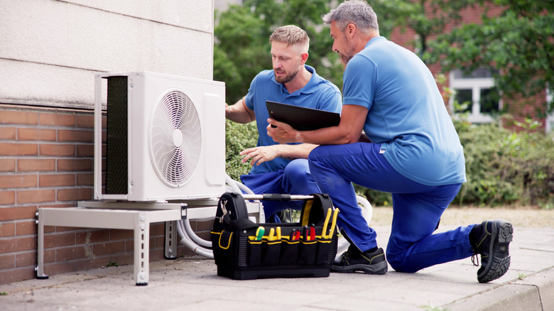 Two men kneeling next to an AC unit outside with tools and a clipboard
