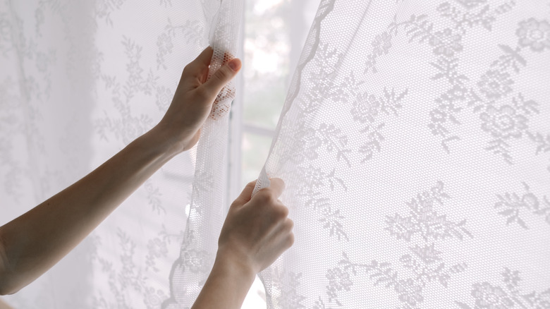 A woman closing white lace curtains in a bedroom