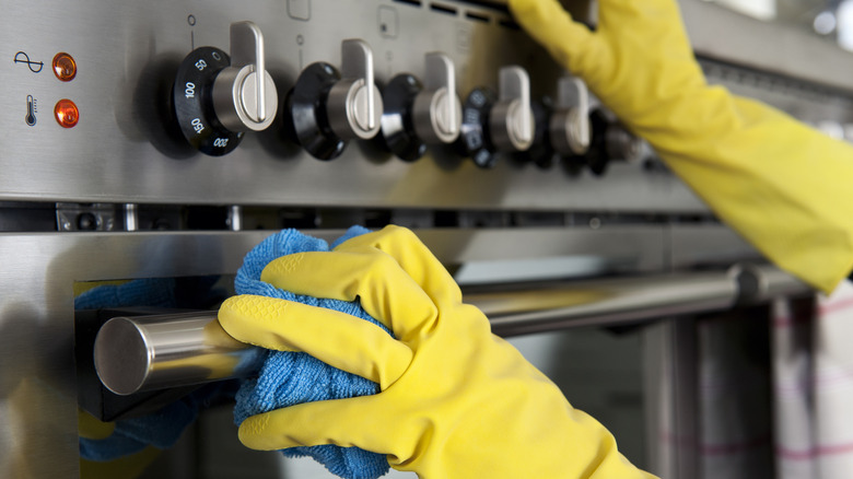 A person wearing yellow gloves wipes a stainless steel oven door with a blue cloth.