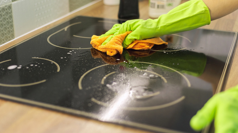 Closeup of a black stovetop being cleaned by a set of hands in green gloves using a microfiber cloth