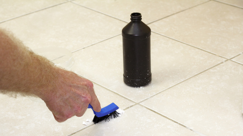 Hand using a blue brush to scrub floor grout sprayed with hydrogen peroxide and a white powder