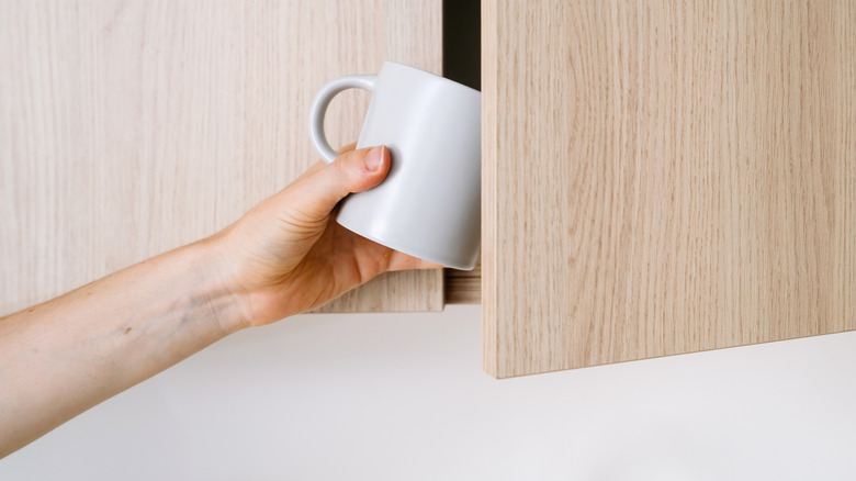 A person grabbing a mug out of a light wood cabinet