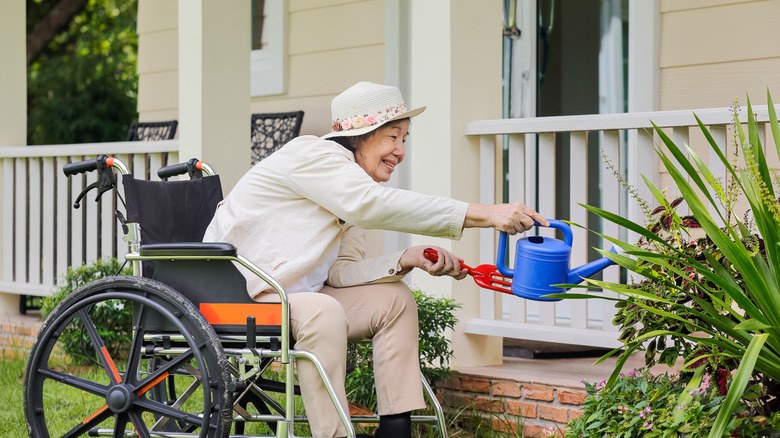 A gardener in a wheelchair stretches to water plants near her porch