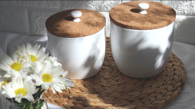 Two white flower pots with DIY lids on top made from IKEA cork trivets, sitting on a counter next to white flowers