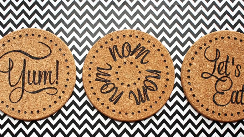 Three HEAT trivets with kitchen-themed designs and phrases burnt into them on a zigzag background