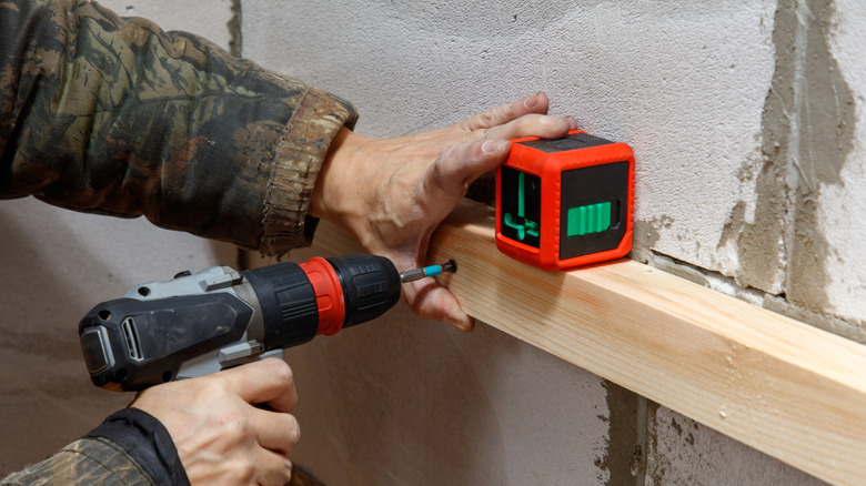 Person holding laser level while drilling into wooden plank