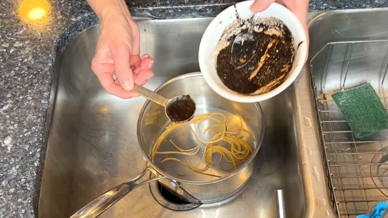 Person spooning used coffee grounds into stainless steel pot in sink