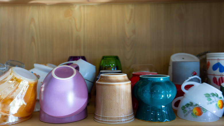 A selection of different sized mugs in different colours sitting on a shelf in a cupboard.