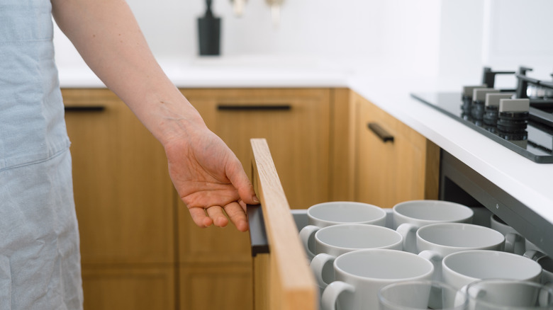 Coffee cups stacked in a drawer with a woman's hand opening the drawer.