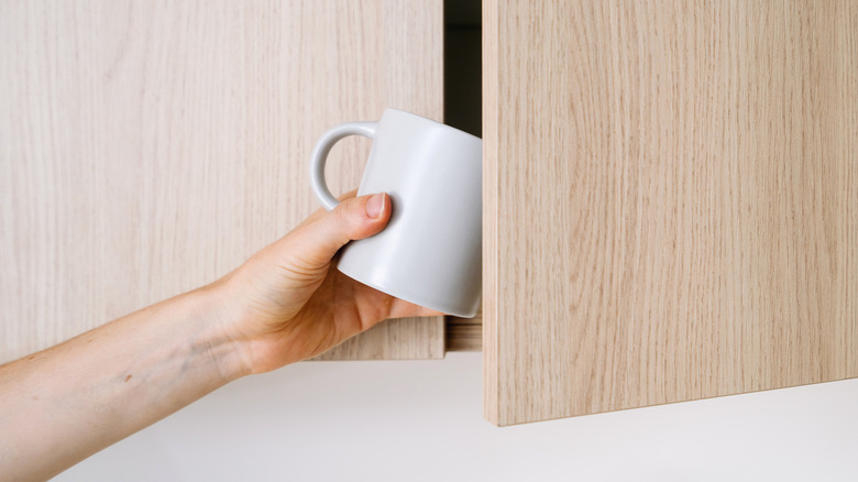 A person grabbing a mug out of a light wood cabinet