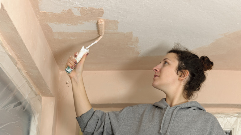A woman uses a small roller to color drench the ceiling of a room using a peachy color