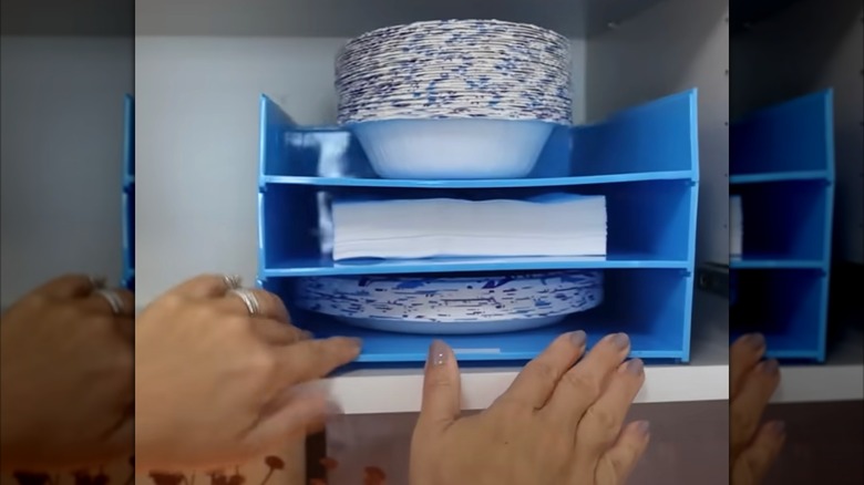 Woman's hands sliding a stack of blue file organizers filled with disposable plates and napkins into kitchen cabinet