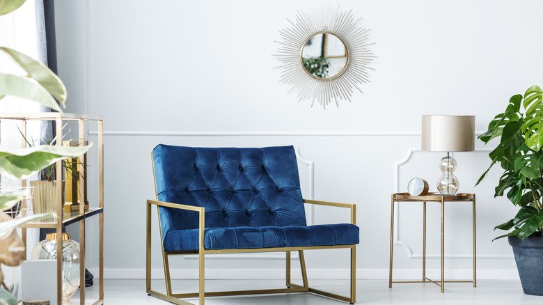 A living room adorned with a blue upholstered chair with a gold metal frame, a gold metal sidetable, a chrome clock, and a silver mirror