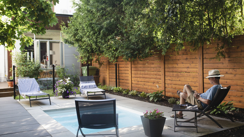A man relaxes in a fenced-in yard while reading a book.