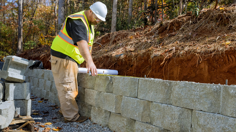 Man measuring a stacked concrete block retaining wall with extra blocks sitting behind him
