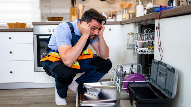 A man crouched over the dishwasher, looking bummed, with various tools around