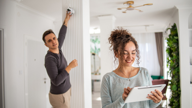 A woman holds a tablet while a man in the background installs a security camera inside a home.