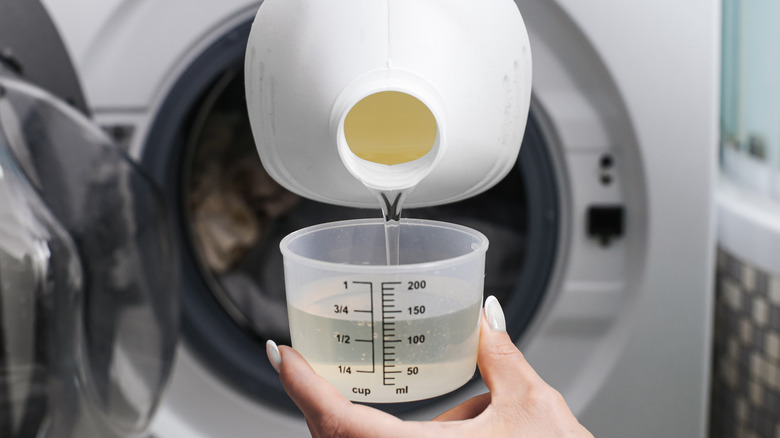 A person pouring clear liquid laundry detergent into a measuring cup in front of a washer