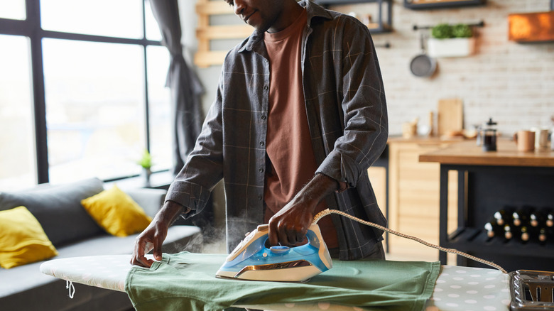 A man irons a tee shirt on an ironing board in a kitchen.