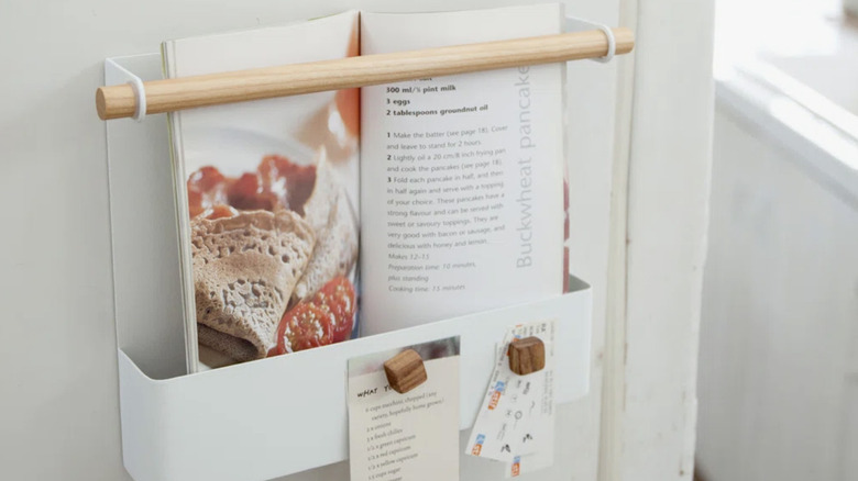 A magnetic book rack on the side of a refrigerator displaying a recipe book.