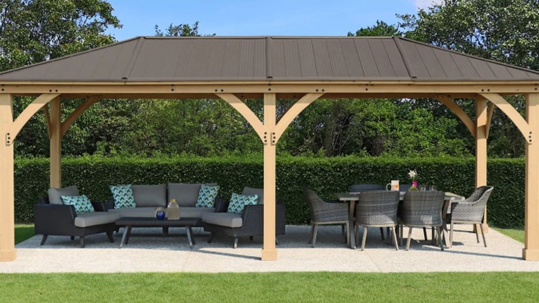 A Yardistry gazebo stands on a concrete slab in a yard and is furnished with seating and a table.