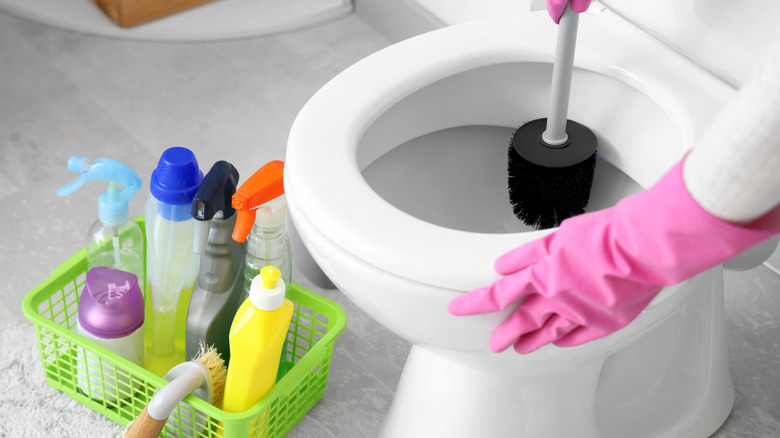 person cleaning toilet with pink gloves, toilet scrubber, and various cleaning products