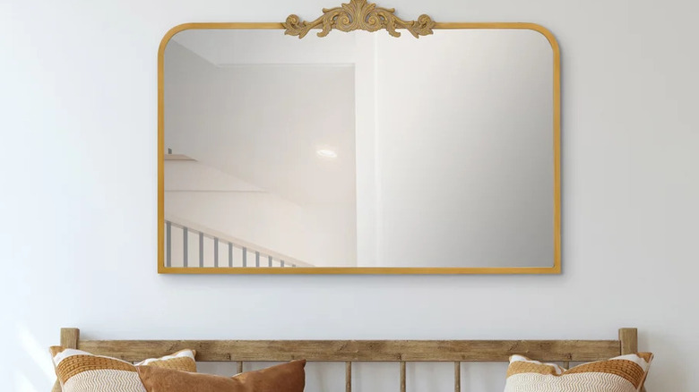 An antique-style mirror with a gold-finished frame hung on a white wall, with a bench sitting below