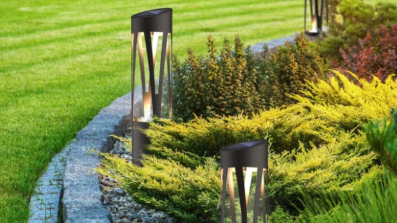 Three solar-powered lighting bollards arranged around a round foliage garden bed with a stone trim, with a grass lawn to the left