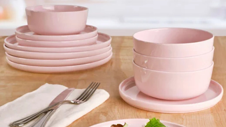Light pink dishware, including round plates with bowls stacked on top of them, on a light wood table with utensils on a napkin nearby