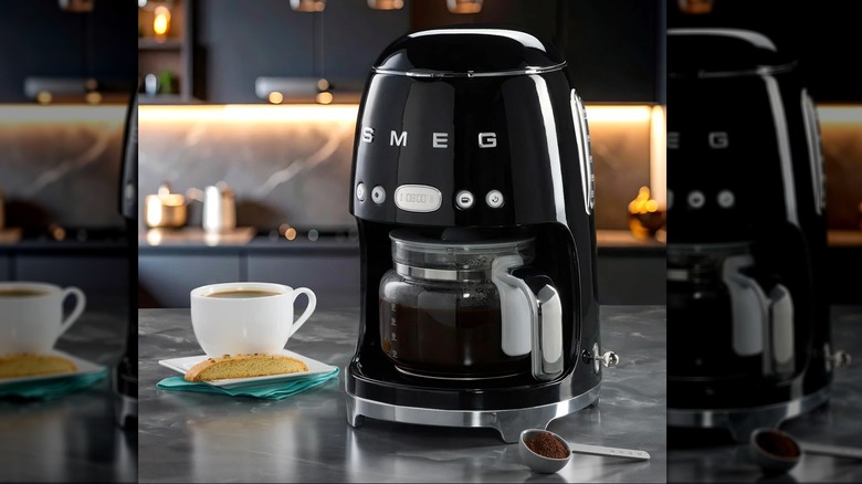 A retro-style drip coffee machine with a shiny back finish on a kitchen counter next to a cup of coffee, biscotti, and a spoonful of coffee grounds
