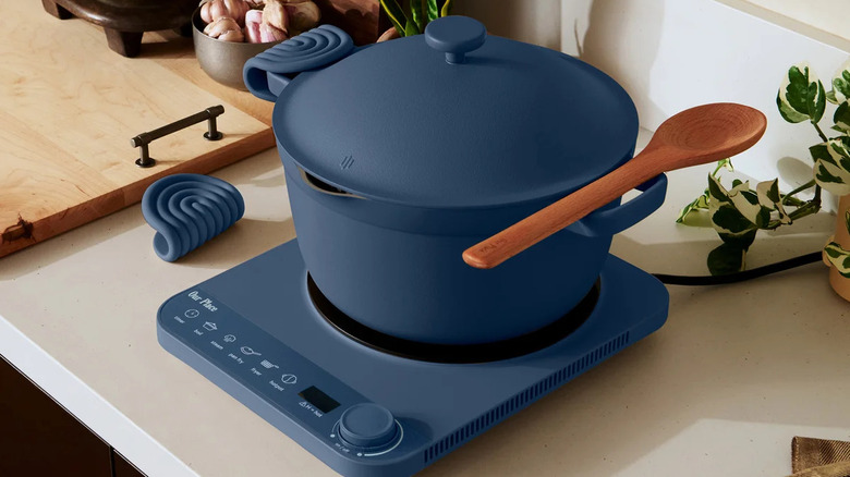 A blue Our Place induction plate and Perfect Pot on a counter with the hot pads and a wooden spoon with a cutting board and plants around it
