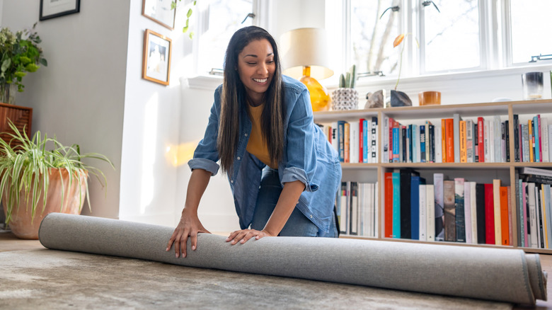 A person unrolling a rug on the floor with plants and a bookshelf in the background
