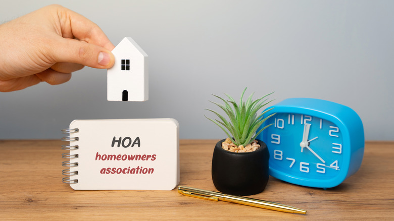 Hand holding a small model home over a wood desk with a homewoner association sign