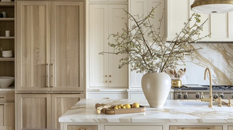 Ivory countertops with tall white oak cabinets and a large vase with a twig arrangement in it. There are also gold taps and the edge of a stainless steel oven in the right of the shot.