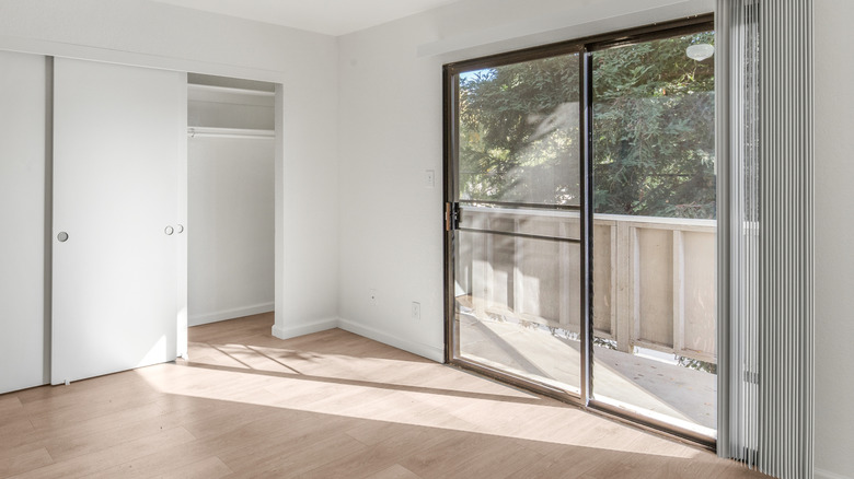 A sliding glass door with coverings opened with direct light streaming onto the floor and part of the wall