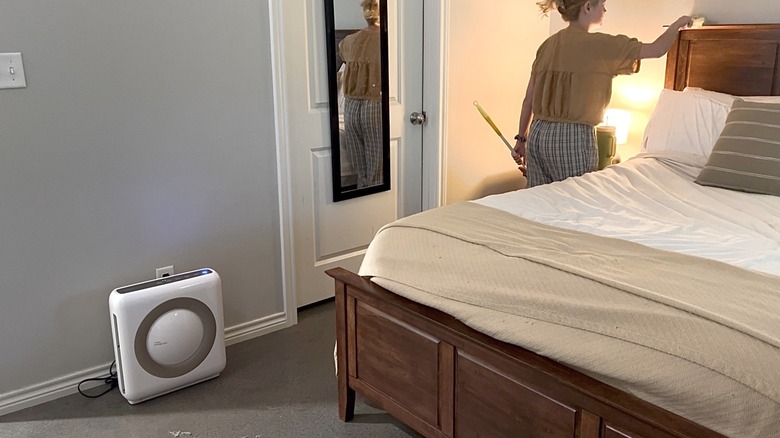 Person dusting bedroom furniture with a Coway Airmega Mighty AP-1512HH air purifier nearby.