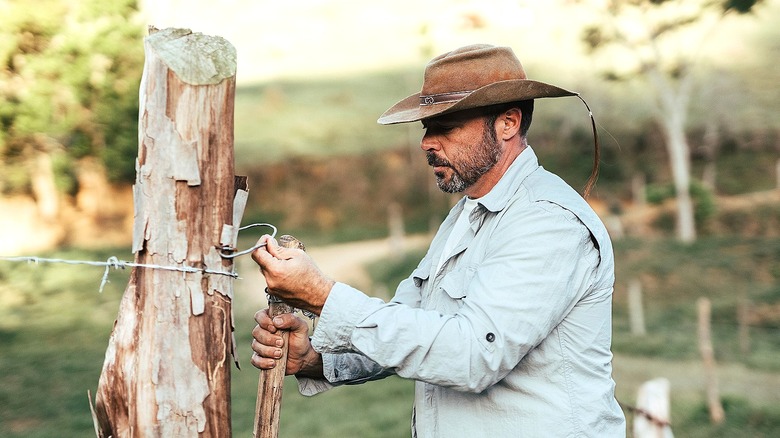 A person wearing a cowboy hat works on a piece of wire and wood next to a large wooden post
