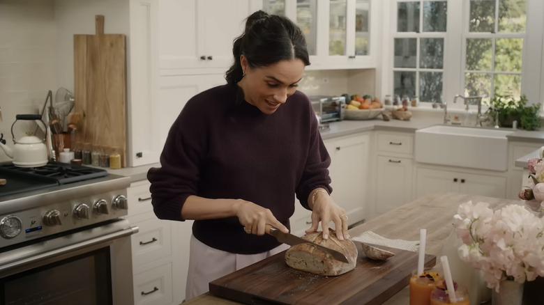 Meghan Markle cuts a loaf of homemade bread on a large wooden cutting board