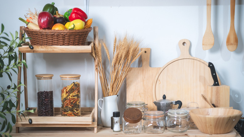 Fruit-filled woven basket, wooden cutting boards, and wooden utensils against a kitchen wall