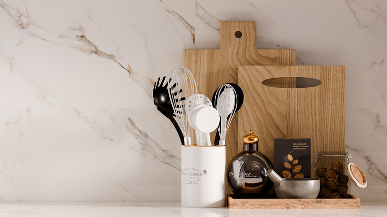 Wooden cutting boards, utensil-filled crock, and tray with kitchen staples on a white marbled counter