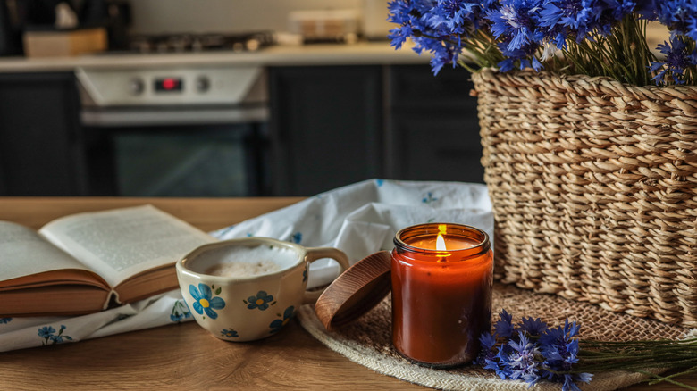 A lit candle in amber jar, ceramic mug with coffee, and flowers in a wicker basket on a wooden kitchen island