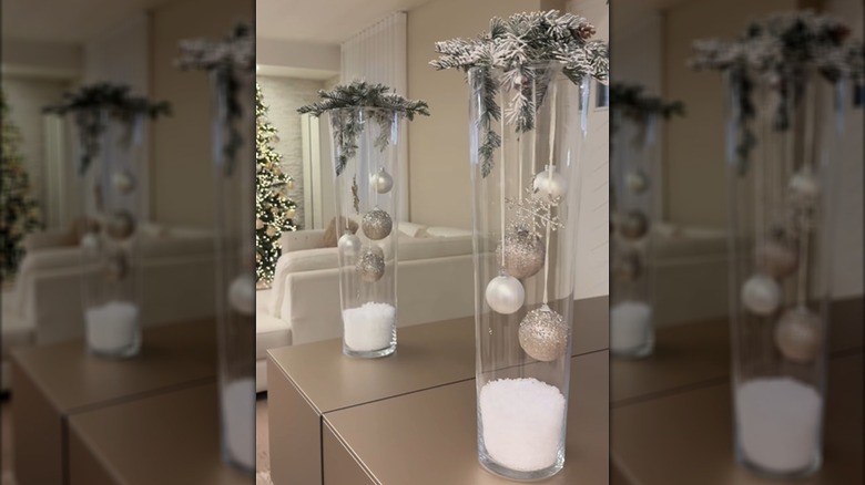 A winter decoration on a table is made of a vase, ornaments, and fake greenery and snow.