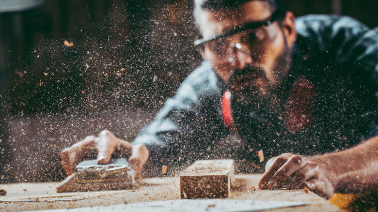 A carpenter in his workshop sands a piece of wood as the shavings fly through the air.