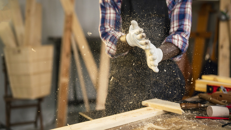A carpenter finishes the day's work by dusting his gloved hands as wood dust flies through the air.